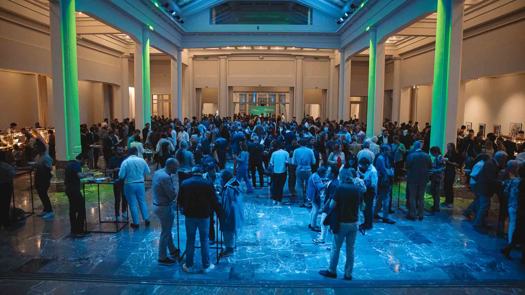 Een grote witte zaal met donkermarmeren vloer die vol met mensen staat.