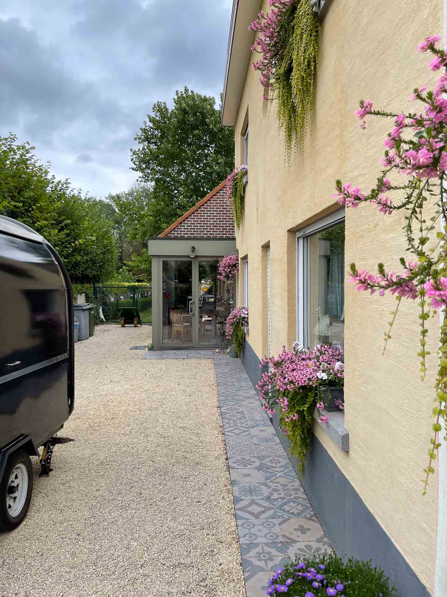 Zijkant van lichtgeel huis met lichtgele kiezelsteentjes en roze bloemen op de vensterbanken.